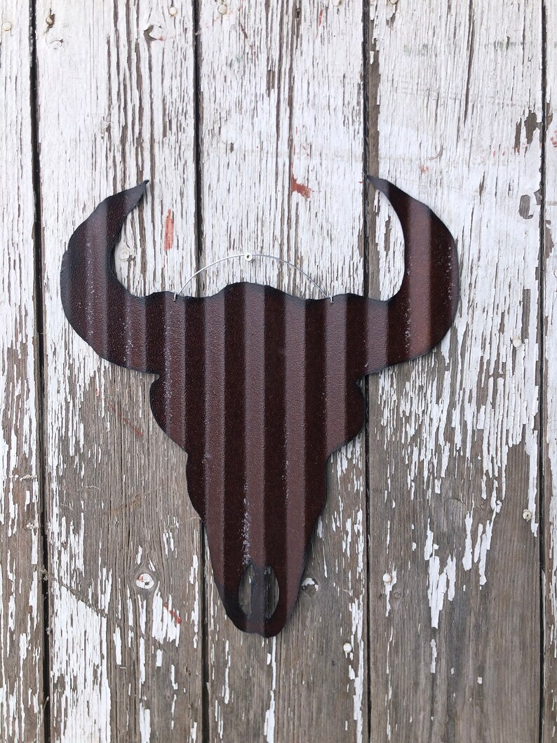 May include: A rustic brown metal bull skull silhouette with a distressed finish. The skull is hanging on a weathered white wooden wall.
