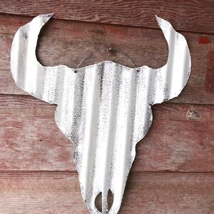 Op de afbeelding: Witte metalen stieren schedel wanddecoratie met een gegolfde textuur. De schedel hangt aan een houten muur.