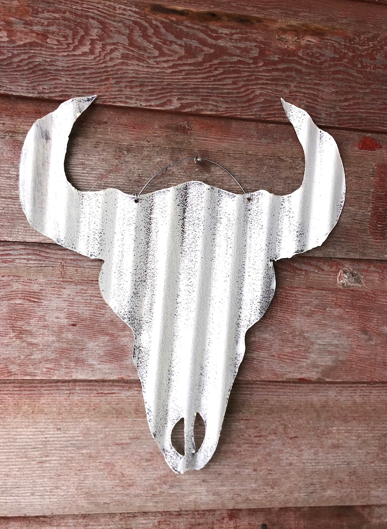 Op de afbeelding: Witte metalen stieren schedel wanddecoratie met een gegolfde textuur. De schedel hangt aan een houten muur.