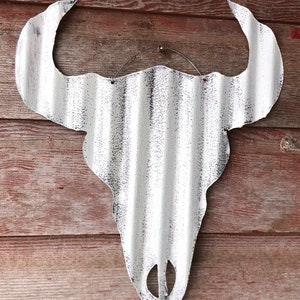 Op de afbeelding: Witte metalen stieren schedel wanddecoratie met een verweerde afwerking. De schedel is gemaakt van gegolfd metaal en heeft een rustieke uitstraling.