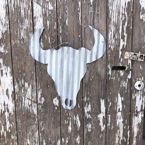 May include: A white metal bull skull silhouette mounted on a weathered wooden door with white paint peeling off.
