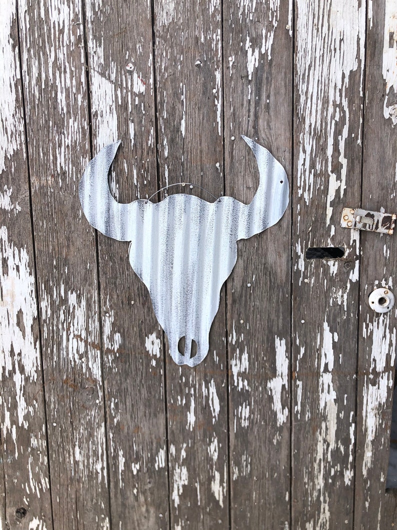 May include: A white metal bull skull silhouette mounted on a weathered wooden door with white paint peeling off.