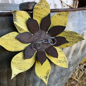 K&ouml;nnte beinhalten: Eine gelbe und braune Metallsonnenblume mit einer abgenutzten Oberfl&auml;che. Die Sonnenblume ist an einem Metallbeh&auml;lter mit rustikalem Aussehen befestigt.