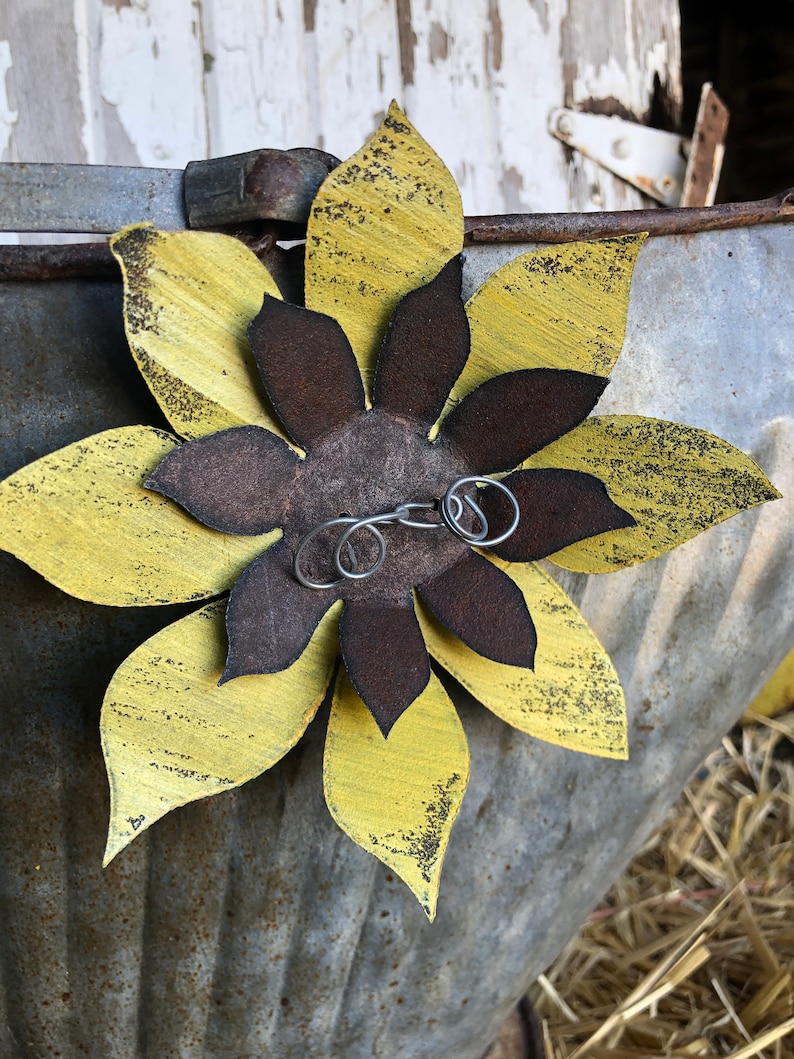 K&ouml;nnte beinhalten: Eine gelbe und braune Metallsonnenblume mit einer abgenutzten Oberfl&auml;che. Die Sonnenblume ist an einem Metallbeh&auml;lter mit rustikalem Aussehen befestigt.