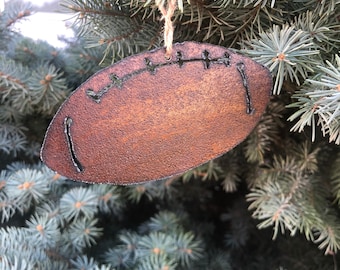 May include: A rustic brown metal football ornament with black stitching details. The ornament is hanging from a twine string on a green pine tree branch.