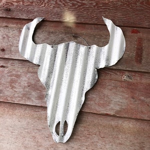 Op de afbeelding: Witte metalen stieren schedel met een gegolfde textuur, hangend aan een houten muur. De schedel is verweerd met een zwarte verflaag.