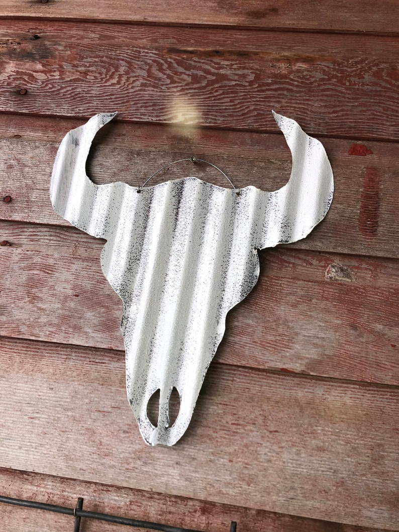 Op de afbeelding: Witte metalen stieren schedel met een gegolfde textuur, hangend aan een houten muur. De schedel is verweerd met een zwarte verflaag.