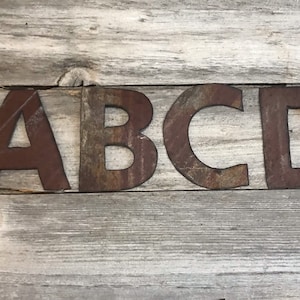 May include: Rustic metal letters spelling out "ABCD" in a brown, rusty color. The letters are arranged in a row on a light wood background.
