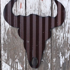 May include: A dark brown metal bull skull silhouette wall decor with a rustic look. The skull is hanging on a white wooden wall with peeling paint.