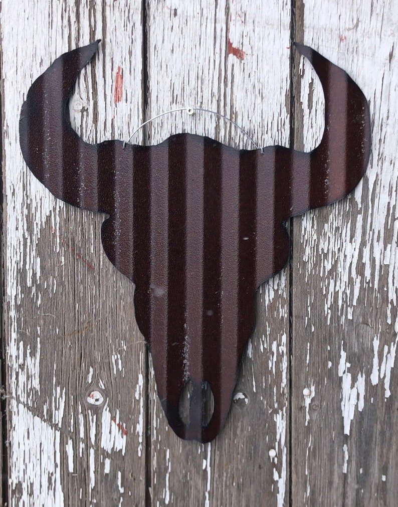 May include: A dark brown metal bull skull silhouette wall decor with a rustic look. The skull is hanging on a white wooden wall with peeling paint.