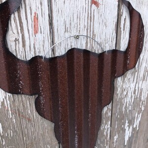 May include: A rusty brown metal bull skull silhouette with a corrugated texture. The skull is hanging from a wire loop.