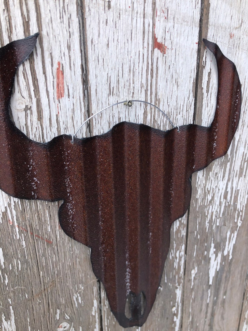 May include: A rusty brown metal bull skull silhouette with a corrugated texture. The skull is hanging from a wire loop.