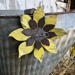 K&ouml;nnte beinhalten: Eine gelbe und braune Metallsonnenblume mit einer Drahtschlaufe, die an der Mitte befestigt ist. Die Sonnenblume ist an einem verzinkten Metallbeh&auml;lter befestigt.