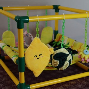 Puede incluir: Un corral de juego amarillo y verde para animales pequeños con un juguete de peluche con forma de limón, una pelota verde y amarilla y un juguete con forma de flor amarilla y rosa que cuelga de la parte superior.