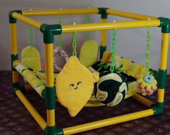 May include: A yellow and green playpen for small animals with a lemon-shaped plush toy, a green and yellow ball, and a yellow and pink flower-shaped toy hanging from the top.