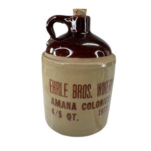 May include: A vintage stoneware jug with a dark brown glazed top and handle, and a tan body. The jug has a cork stopper and features the text "Ehrle Bros. Winery Amana Colonies 4/5 QT. 1973".