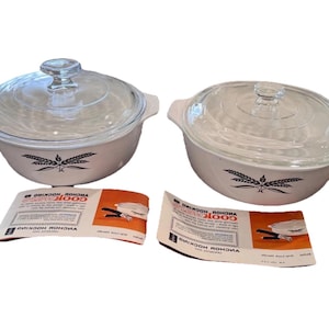 May include: Two white casserole dishes with clear glass lids. Each dish has a black wheat design and side handles. The lids have a knob handle. Two instruction sheets are in front of the dishes.