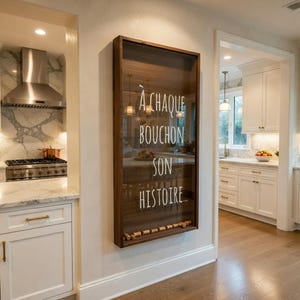 May include: A wall-mounted wine cork display case with a dark brown wooden frame and a glass front. The text "À CHAQUE BOUCHON SON HISTOIRE..." is written in white. A row of corks is visible at the bottom of the case.