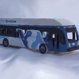 May include: A miniature blue and white bus model with the text "ZERO EMISSION" on the roof. The bus features the letter "V" in a light blue design on the side and the letters "VA" on the front.