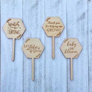 May include: Four wooden garden stakes with different messages: "Watch me grow", "Let love grow", "Thank you for helping me grow!" and "Baby in bloom".