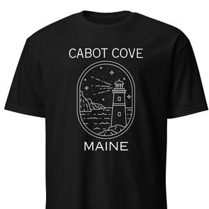 Può includere: T-shirt nero con un design grafico bianco di un faro, stelle e onde. Il testo "CABOT COVE MAINE" è stampato sulla maglietta.