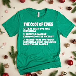 Op de afbeelding: Een groen t-shirt met de tekst "THE CODE OF ELVES" en drie kerstregels. Het shirt is omgeven door kerstversieringen, dennennaalden en dennenappels.