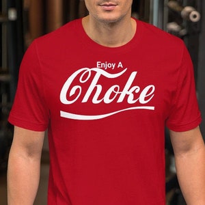 Op de afbeelding: Rood T-shirt met korte mouwen met een witte afbeelding met de tekst "Enjoy A Choke" in een gestileerd lettertype.