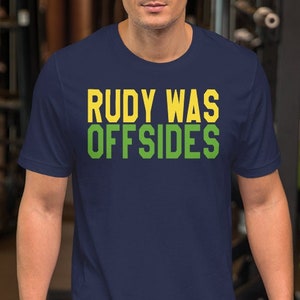 Op de afbeelding: Een marineblauw t-shirt met de tekst "Rudy Was Offsides" in gele en groene letters.