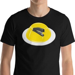 May include: A black t-shirt featuring a yellow jelly mould with a black stapler on top. The jelly mould is on a white plate.
