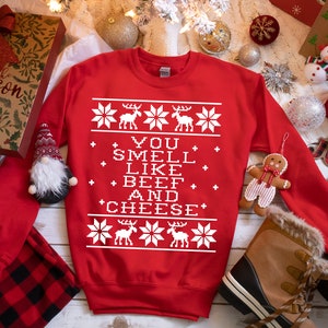 May include: A red sweatshirt with a white, pixelated design featuring reindeer and snowflakes. The text "YOU SMELL LIKE BEEF AND CHEESE" is displayed in the center. The sweatshirt is surrounded by Christmas decorations.