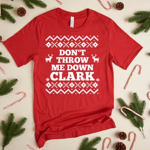 Puede incluir: Una camiseta roja con el texto blanco "DON'T THROW ME DOWN CLARK". La camiseta presenta un diseño festivo con motivos de renos y copos de nieve, ideal para la temporada navideña. La camiseta está rodeada de bastones de caramelo, piñas y ramas de pino.