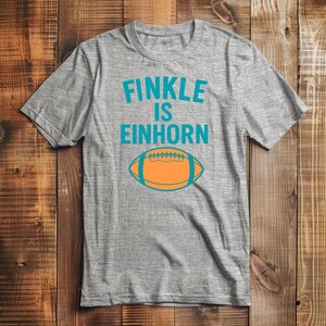 Pu&ograve; includere: Una maglietta grigio melange con la scritta turchese "FINKLE IS EINHORN" sopra una grafica di football americano arancione e turchese. La maglietta ha maniche corte e girocollo.