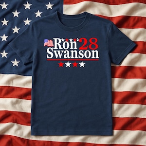 May include: A navy blue t-shirt with the text "Ron '28 Swanson" in white and red, featuring an American flag graphic. The design includes red and white stars. The shirt is displayed against an American flag background.