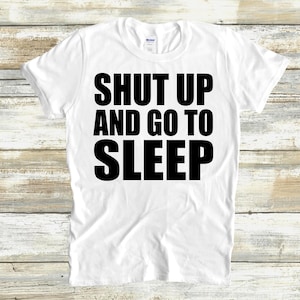 Puede incluir: Camiseta blanca con la frase "SHUT UP AND GO TO SLEEP" impresa en letras negras y grandes. La camiseta tiene cuello redondo y está hecha de una tela suave.
