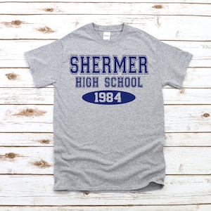 Può includere: Maglietta grigia con testo blu che recita "Shermer High School 1984".