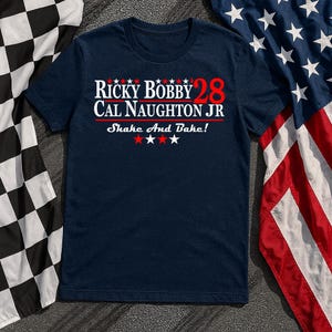 Könnte beinhalten: Marineblaues T-Shirt mit dem Text "Ricky Bobby 28 Cal Naughton Jr Shake And Bake!" in Weiß und Rot. Das Shirt wird mit einer amerikanischen Flagge und einer Zielflagge im Hintergrund präsentiert.