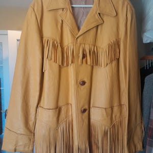 May include: A tan leather jacket with fringe detailing across the chest and pockets. The jacket has a collared neckline, two brown buttons, and a label inside the collar. The jacket is hanging on a hanger.