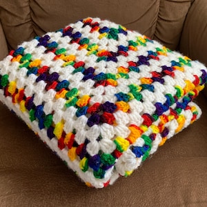 May include: A folded, handmade crochet blanket. The blanket features a white base with colorful squares of red, orange, yellow, green, blue, and purple. The blanket is folded and resting on a brown surface.