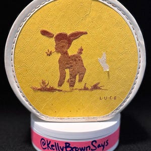 May include: A round, vintage-style decorative piece featuring a brown deer silhouette on a yellow background with a textured pattern. The piece is framed in white and has the text "L.U.C.E." and "@KellyBrownSays".