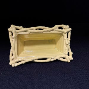May include: A rectangular, light yellow ceramic planter with a decorative, branch-like border. The interior is a slightly darker shade of yellow. The planter is empty and sits on a dark blue surface.