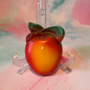 May include: A ceramic apple figurine with red and yellow gradients, green leaves, and a clear plastic stand. The apple is glossy and detailed, showcasing a realistic design. The background is a soft blend of pink and green.