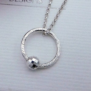May include: A silver necklace with a textured circle pendant and a small, faceted silver bead. The necklace is on a white background.