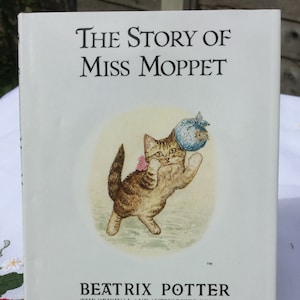 May include: A vintage book cover featuring a brown and white kitten playing with a blue ball. The title of the book is "The Story of Miss Moppet" by Beatrix Potter. The book is part of the original and authorized edition with new color reproductions published by F. Warne & Co.