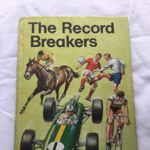 May include: A yellow book cover with the title "The Record Breakers" in black text. The cover features illustrations of a racehorse, a soccer game, a race car, and a cyclist. The book is part of the "Ladybird Key Words Easy Readers" series and is labeled "Book One".
