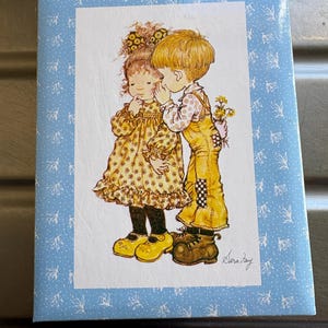 Peut inclure: Une couverture de livre vintage avec une illustration de deux enfants. La fille porte une robe jaune à motifs floraux, et le garçon porte une salopette jaune. Le garçon chuchote à l'oreille de la fille, tenant des fleurs derrière son dos. La couverture a un motif floral bleu.