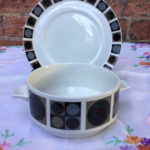 Peut inclure: Un bol en céramique blanche avec un motif géométrique noir et gris. Le bol a deux anses et est posé sur une nappe blanche à broderies florales.