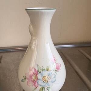 May include: A white ceramic vase with a floral design. The vase features a light blue and pink flower with green leaves. The vase has a handle on one side.