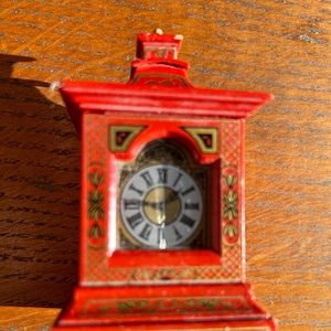 Puede incluir: Un pequeño reloj rojo con detalles dorados y una esfera blanca. El reloj tiene un diseño decorativo en la parte delantera y una parte superior puntiaguda.