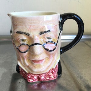 Könnte beinhalten: Eine Keramikkanne in Form eines Männergesichts mit Brille und Fliege. Die Kanne hat einen schwarzen Henkel und eine weiße Glasur mit einer strukturierten Oberfläche.