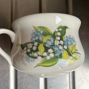May include: A white ceramic teacup with a floral design featuring blue and white flowers and green leaves. The cup has a handle and a rounded shape.
